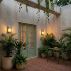 Clérachic upright wall lamps lit amber at dusk beside a sage green French door, with palms, ferns and a small bistro table.