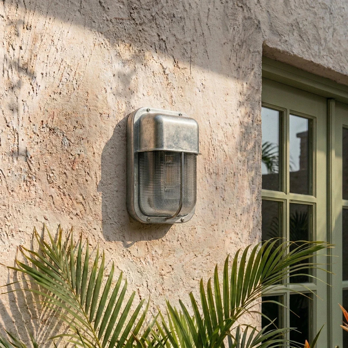 Clérachic upright wall lamp in raw galvanised metal with a ribbed glass shade, on a rough plaster wall beside a green window.
