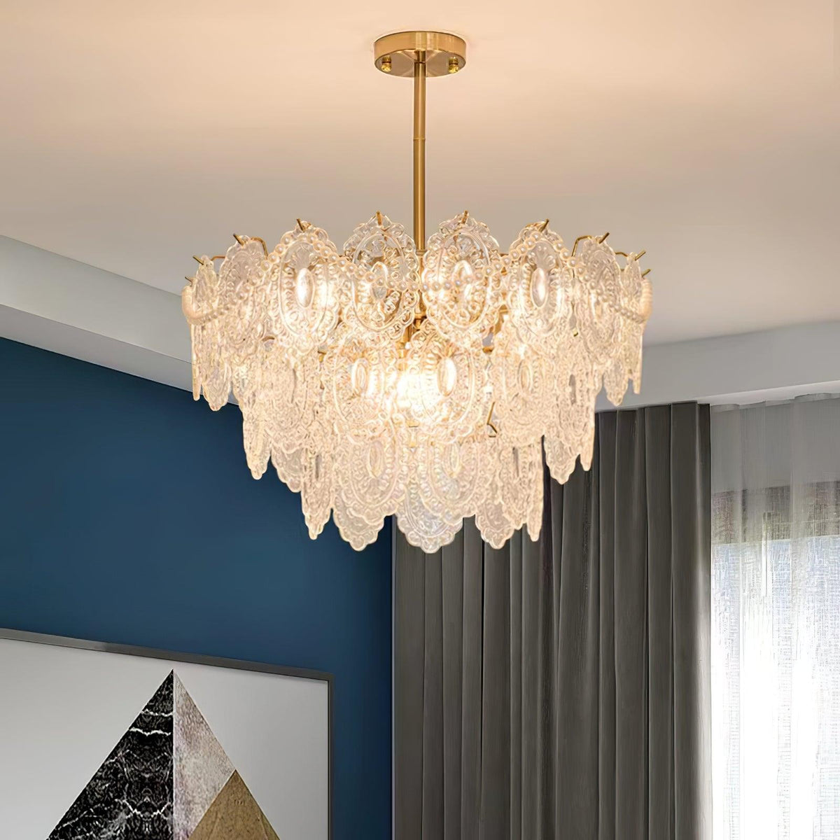 Lit Clérachic glass chandelier with a gold stem, in a room with a deep blue wall and grey curtains.