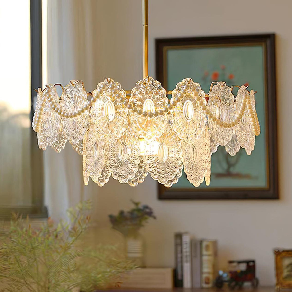 A Clérachic chandelier glowing warm above a sideboard with books, a framed picture and a leafy plant.