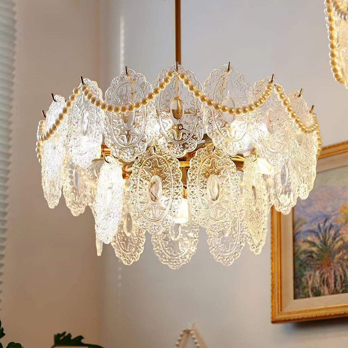 Side view of a Clérachic chandelier, its patterned glass panels and pearl strands lit, beside a gilt-framed painting.