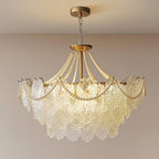 A Clérachic glass and pearl pendant glowing warmly on a gold chain against a soft pink-beige wall, with its gold canopy at the top.