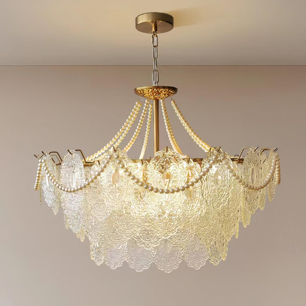 A Clérachic glass and pearl pendant glowing warmly on a gold chain against a soft pink-beige wall, with its gold canopy at the top.