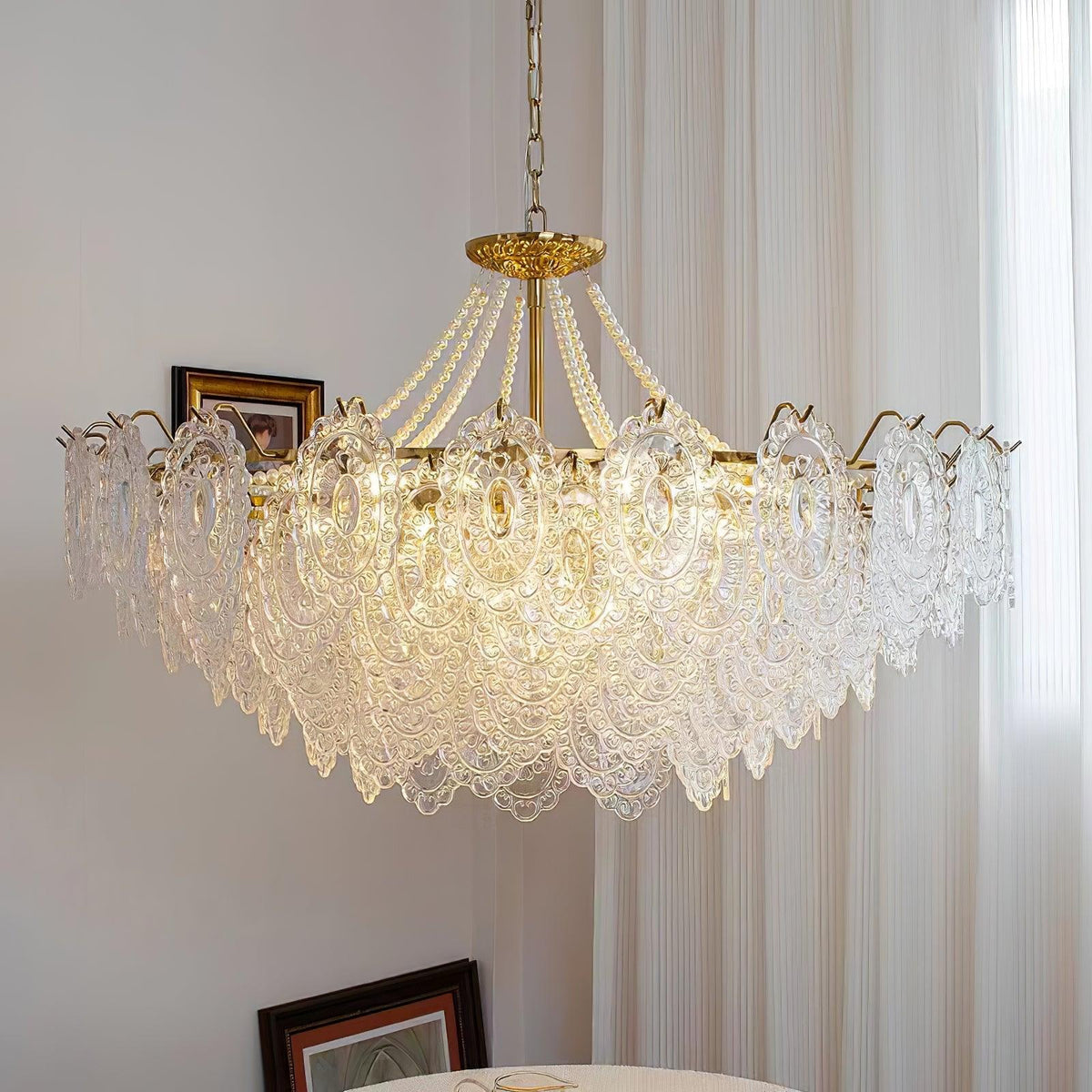 Wide Clérachic pendant light with layered clear glass petals and pearl swags, hanging in front of tall sheer curtains and a framed picture.