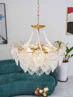 A Clérachic pendant seen from below with its gold frame, pearl swags and glass panels, with a green sofa and a potted plant behind.