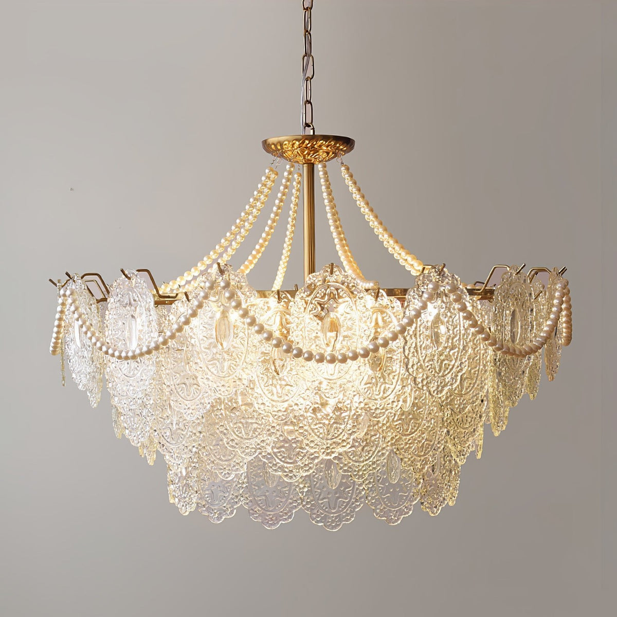 This Clérachic pendant light, lit and hanging on a chain, with glass panels in a stepped shape and pearl swags against a grey wall.