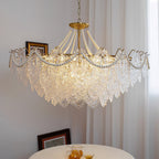 Clérachic pendant with a wide skirt of embossed glass panels and pearl swags, lit beside a framed picture and a sheer curtain, with a table below.