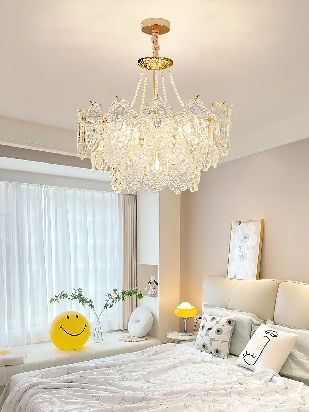 A Clérachic pendant with embossed glass and pearls close to the ceiling in a bedroom with a white bed, a sheer window curtain and a yellow smiley cushion.