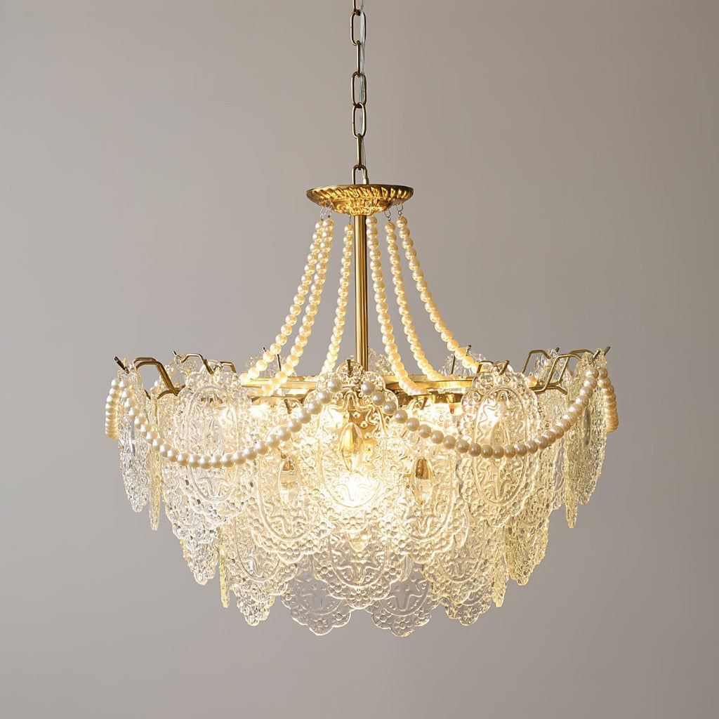 Lit Clérachic pendant with a narrower skirt of embossed glass, pearl swags over a gold frame and a chain, against a grey wall.