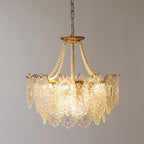 Clérachic pendant with a gold frame and a ring of embossed glass panels, with pearl strands rising to the canopy, lit against a grey wall.