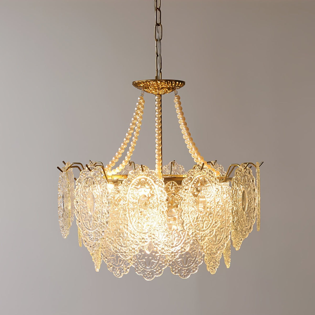 Clérachic pendant with a gold frame and a ring of embossed glass panels, with pearl strands rising to the canopy, lit against a grey wall.