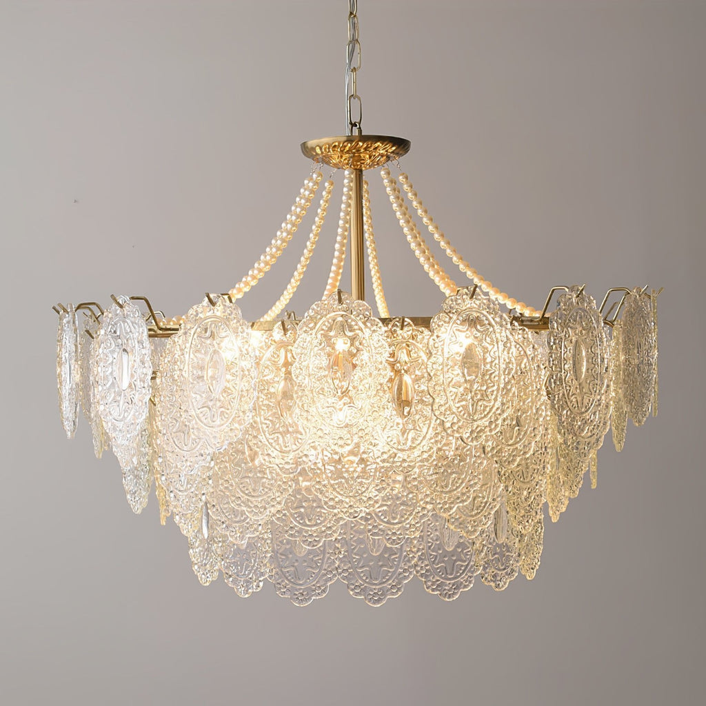 Close view of the wide, lit glass skirt on this Clérachic pendant, with pearl strands rising to its gold canopy on a grey wall.