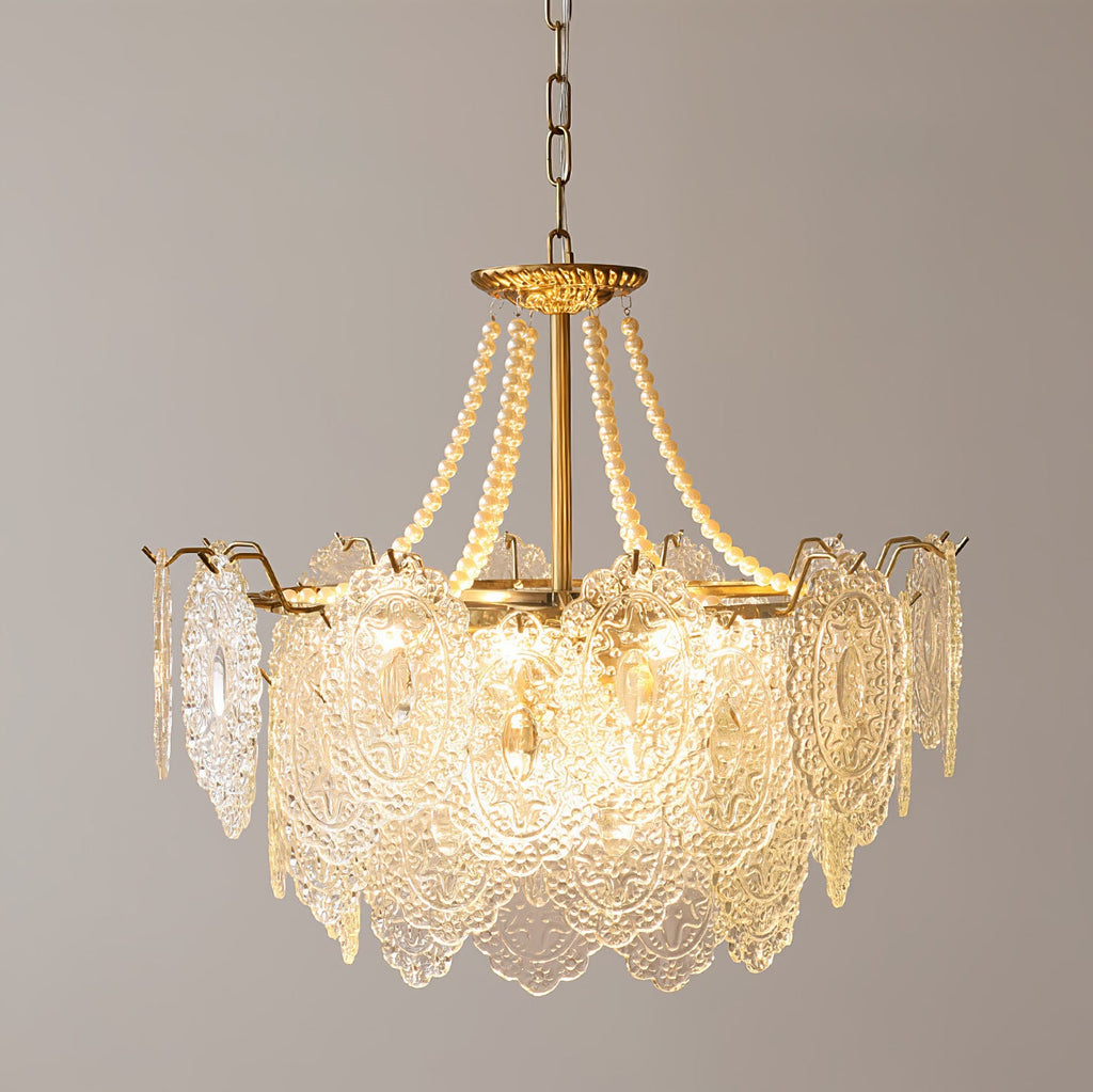 Lit Clérachic pendant with a tall skirt of embossed glass petals and pearl strands on a gold frame, against a grey wall.