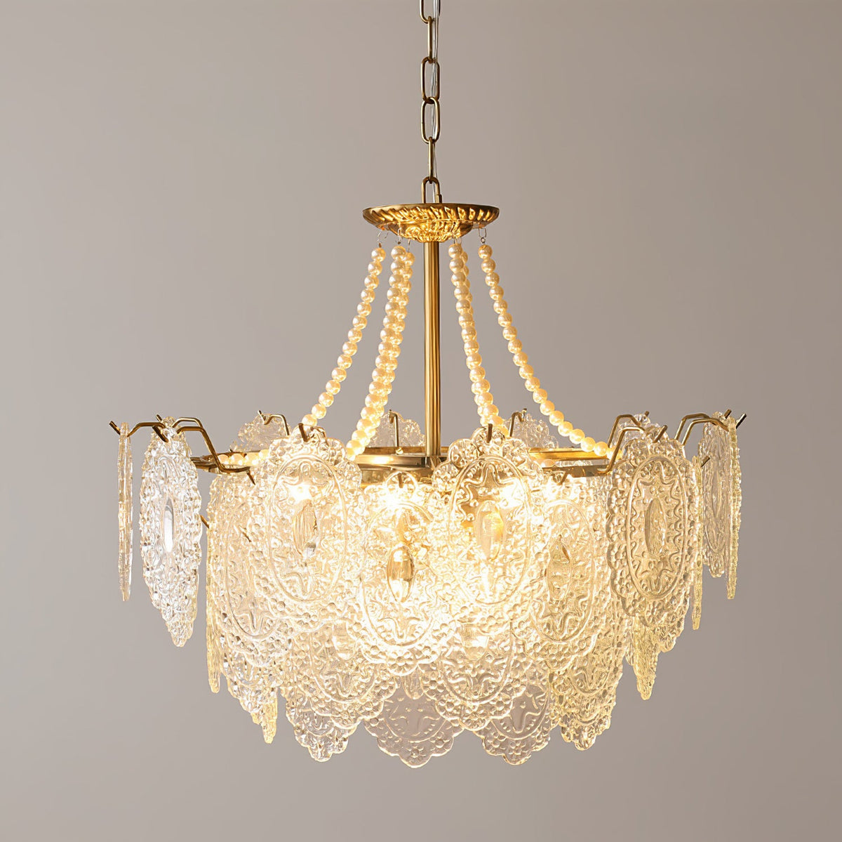 Lit Clérachic pendant with a tall skirt of embossed glass petals and pearl strands on a gold frame, against a grey wall.