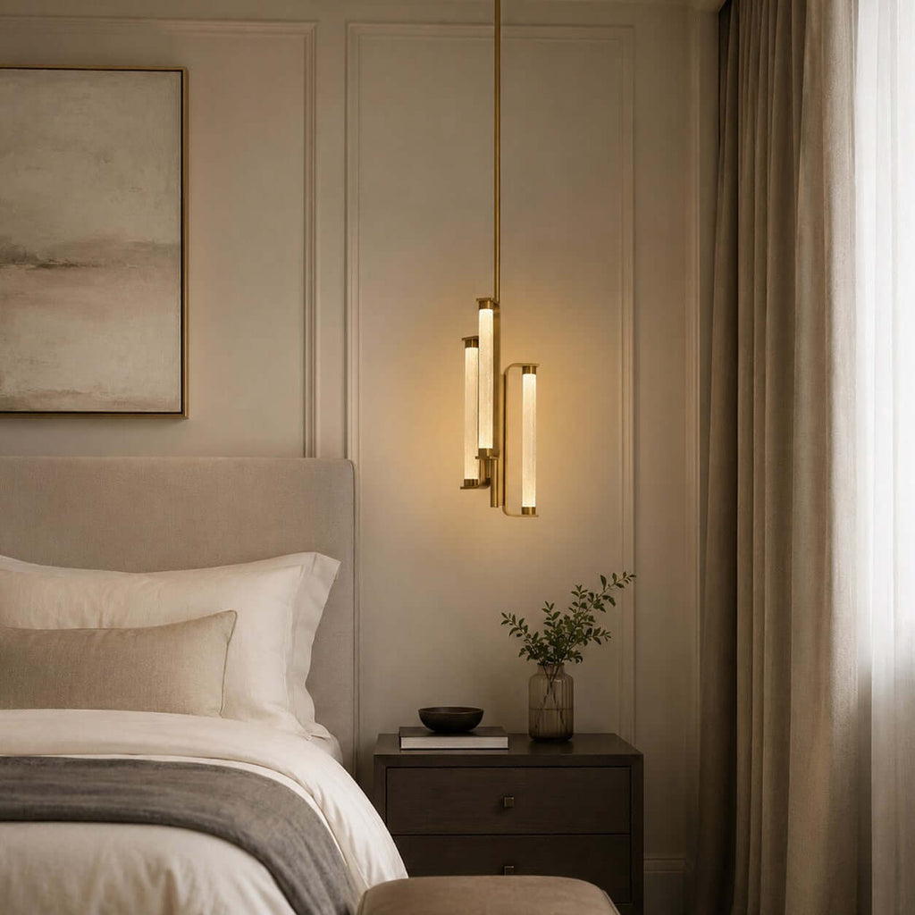 A lit Clérachic pendant with glass tubes in a brass frame hangs beside a bed with a linen headboard, a dark bedside table and a long curtain.