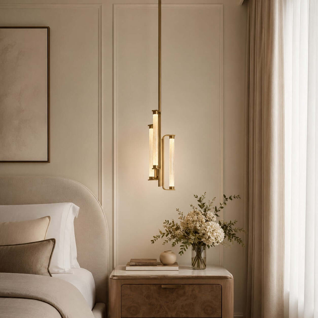 A Clérachic brass and glass pendant glowing beside a curved headboard, above a pale nightstand with a vase of dried flowers.