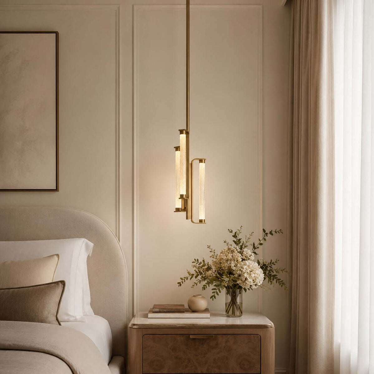 A Clérachic brass and glass pendant glowing beside a curved headboard, above a pale nightstand with a vase of dried flowers.