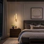 Over a bedside table with a flower vase and a framed print, a Clérachic brass and glass pendant glows warm above a dark upholstered bed.