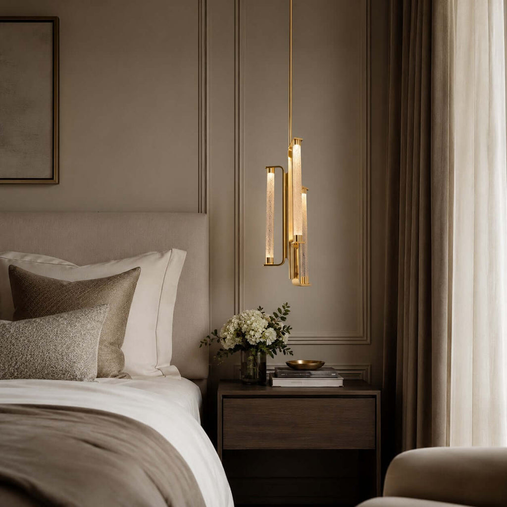 A Clérachic pendant with brass-framed glass tubes hangs close to a bed with a tan headboard, a small tray and a bunch of white flowers.