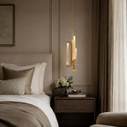 A Clérachic pendant with brass-framed glass tubes hangs close to a bed with a tan headboard, a small tray and a bunch of white flowers.