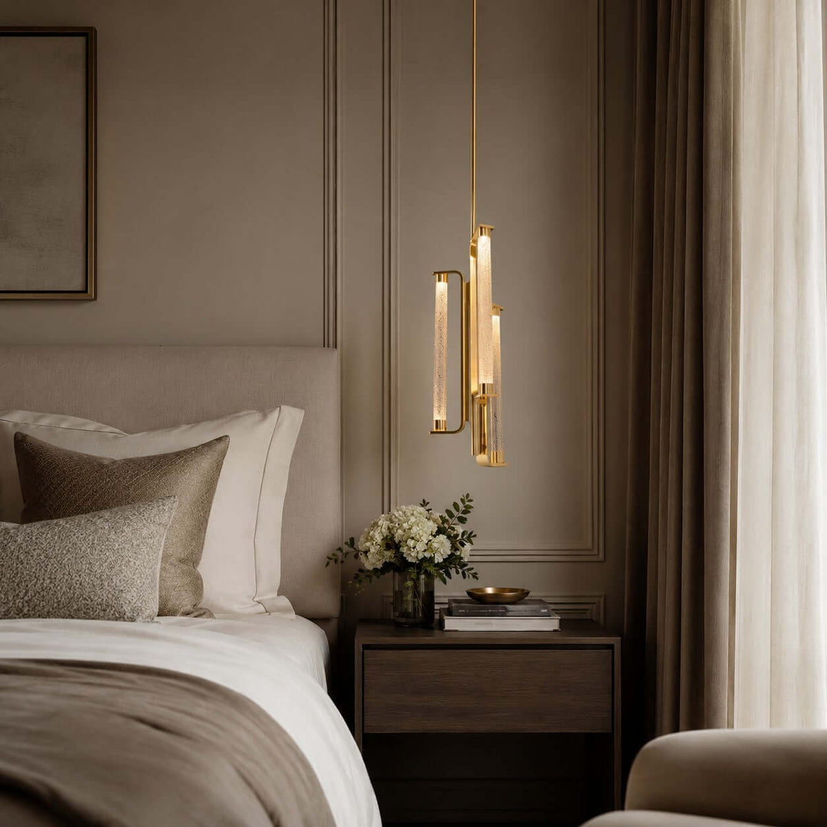 A Clérachic pendant with brass-framed glass tubes hangs close to a bed with a tan headboard, a small tray and a bunch of white flowers.