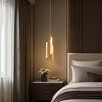 A Clérachic brass pendant lit warm beside a bed with a pale headboard, in front of a dark wood panel and a grey curtain.