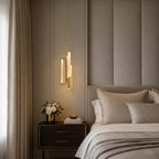 Hung low beside a padded wall panel and a dark bedside chest, a Clérachic brass and glass pendant glows next to a vase of white roses.