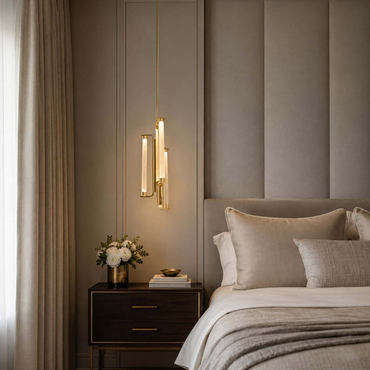 Hung low beside a padded wall panel and a dark bedside chest, a Clérachic brass and glass pendant glows next to a vase of white roses.