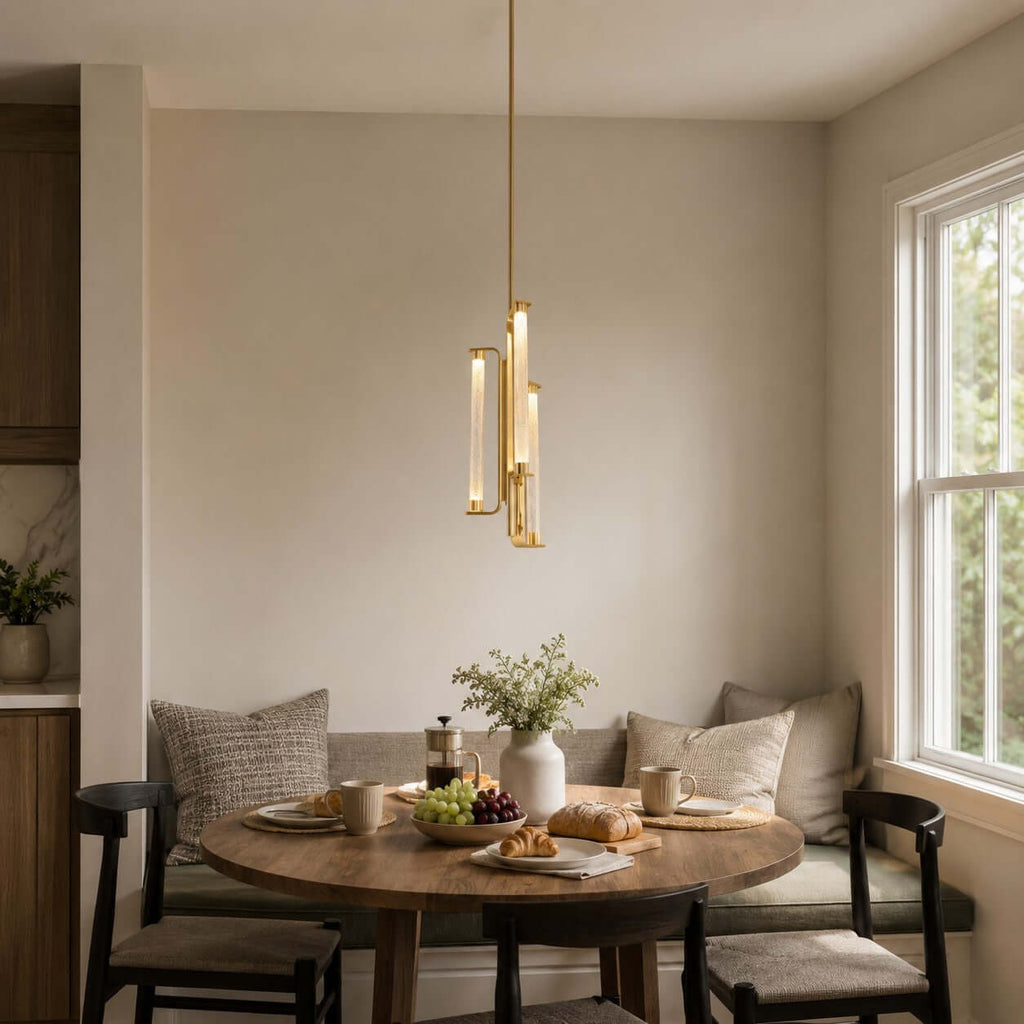 A Clérachic pendant with glass tubes in a brass frame hangs above a small wooden table by a window, in a breakfast corner with a banquette.