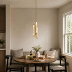 A Clérachic pendant with glass tubes in a brass frame hangs above a small wooden table by a window, in a breakfast corner with a banquette.