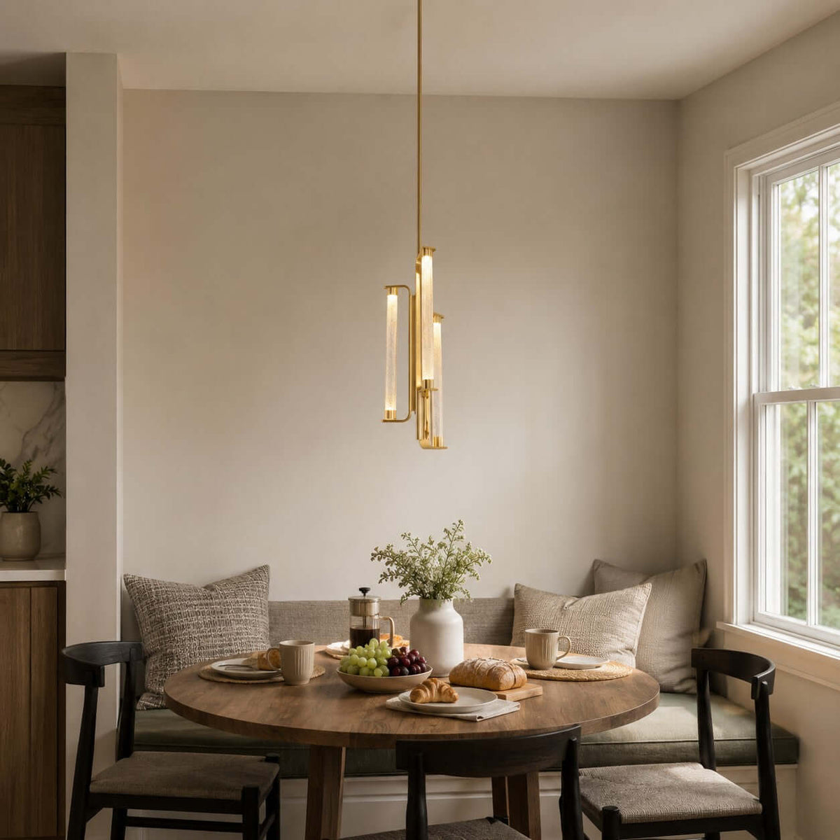 A Clérachic pendant with glass tubes in a brass frame hangs above a small wooden table by a window, in a breakfast corner with a banquette.