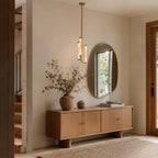 Beside a rounded mirror and a pale wood sideboard, a Clérachic brass pendant lights an entrance hall with a vase of dried stems.