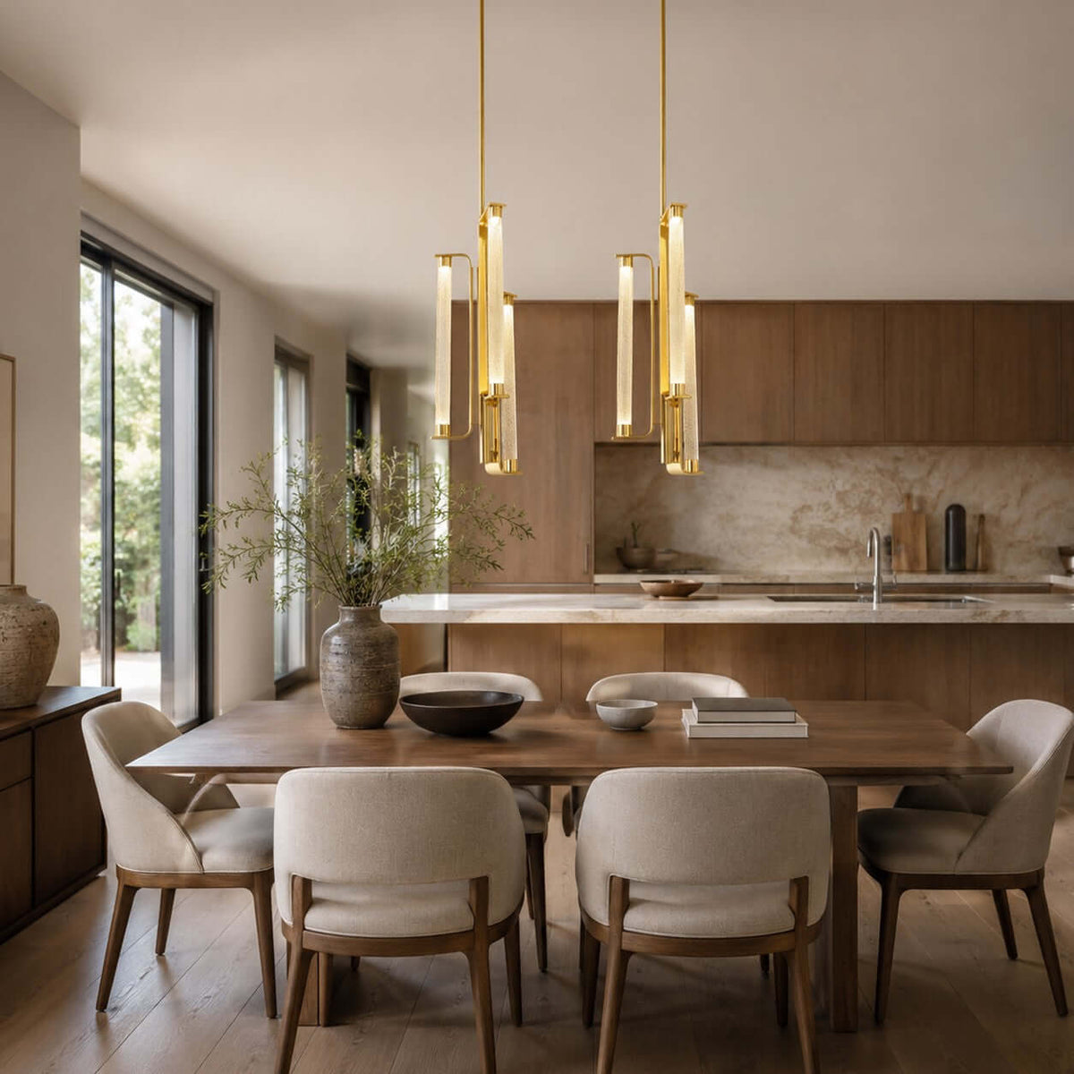 Two Clérachic brass and glass pendants hang side by side over a wooden dining table with linen chairs and an island kitchen behind.