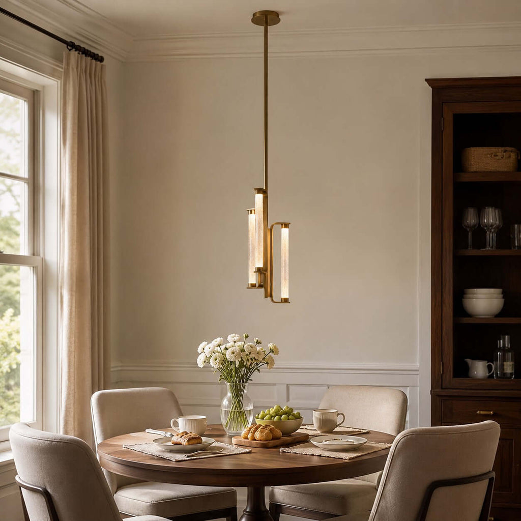 A Clérachic brass pendant hangs above a round dining table with a vase of white flowers, beside a tall window and a glazed dresser.