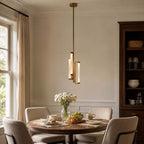 A Clérachic brass pendant hangs above a round dining table with a vase of white flowers, beside a tall window and a glazed dresser.