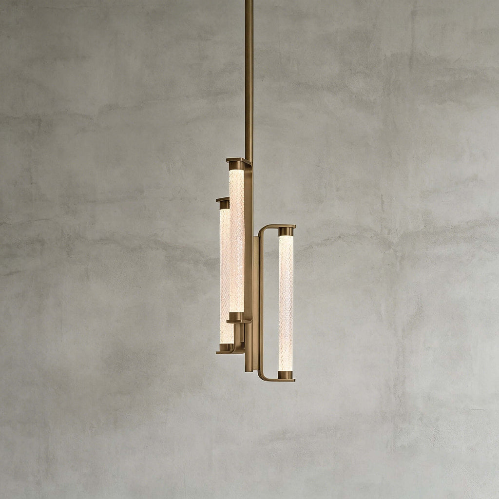This Clérachic pendant on a mid-grey plaster backdrop, showing three glowing textured glass tubes held in a brass frame on a long rod.