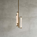 This Clérachic pendant on a mid-grey plaster backdrop, showing three glowing textured glass tubes held in a brass frame on a long rod.