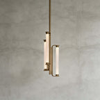 This Clérachic pendant on a mid-grey plaster backdrop, showing three glowing textured glass tubes held in a brass frame on a long rod.