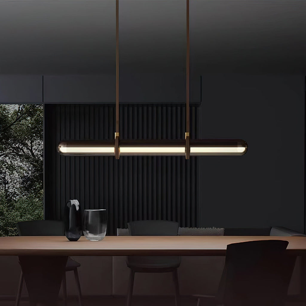 A Clérachic glass tube pendant lit above a dark dining table, hung from two brown leather straps against a dark slatted wall.