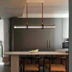 A Clérachic glass tube pendant hung from brown straps over a pale kitchen table, with grey cabinets and leather-seated chairs behind.