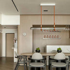A Clérachic glass tube pendant on leather straps over a long dining table in a light wood room, with a small shelf of bottles behind.