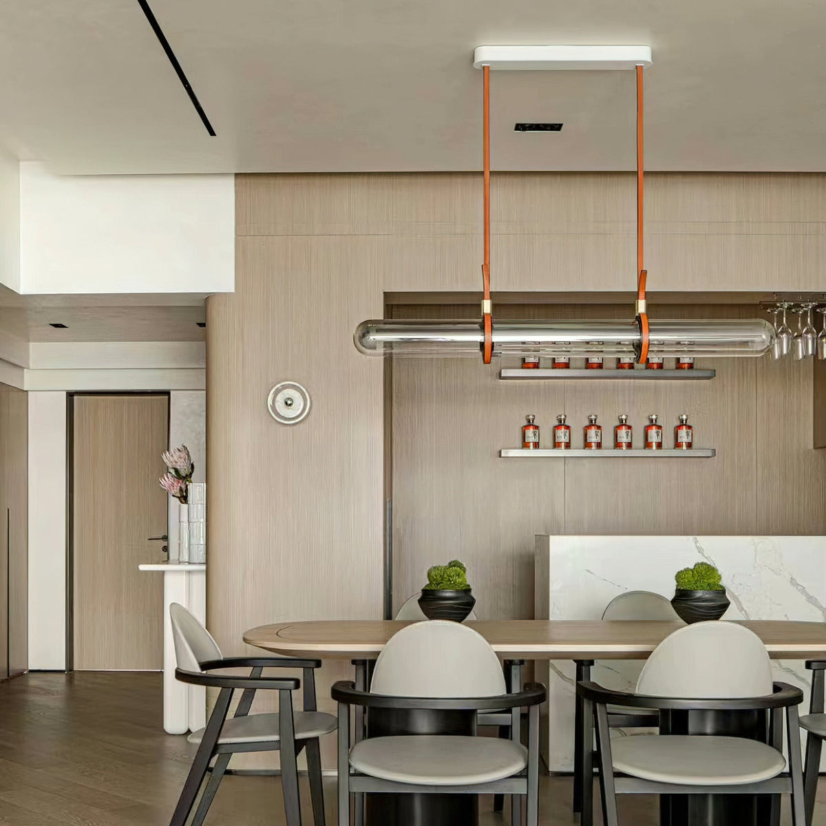A Clérachic glass tube pendant on leather straps over a long dining table in a light wood room, with a small shelf of bottles behind.