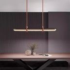 A Clérachic glass tube pendant on two straps against a dark fluted wall, above a wooden table with a vase of dried stems and an open book.