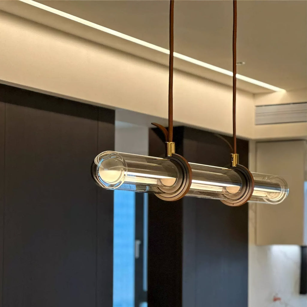 Close view of a Clérachic glass tube pendant, with brown leather loops and brass collars, lit beneath a ceiling with a cove light.