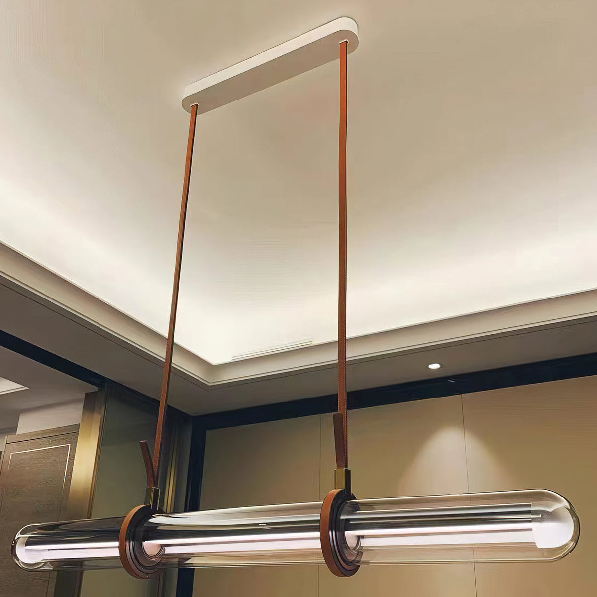 Looking up at a Clérachic glass tube pendant, with long leather straps running to a white ceiling plate beneath a recessed ceiling.