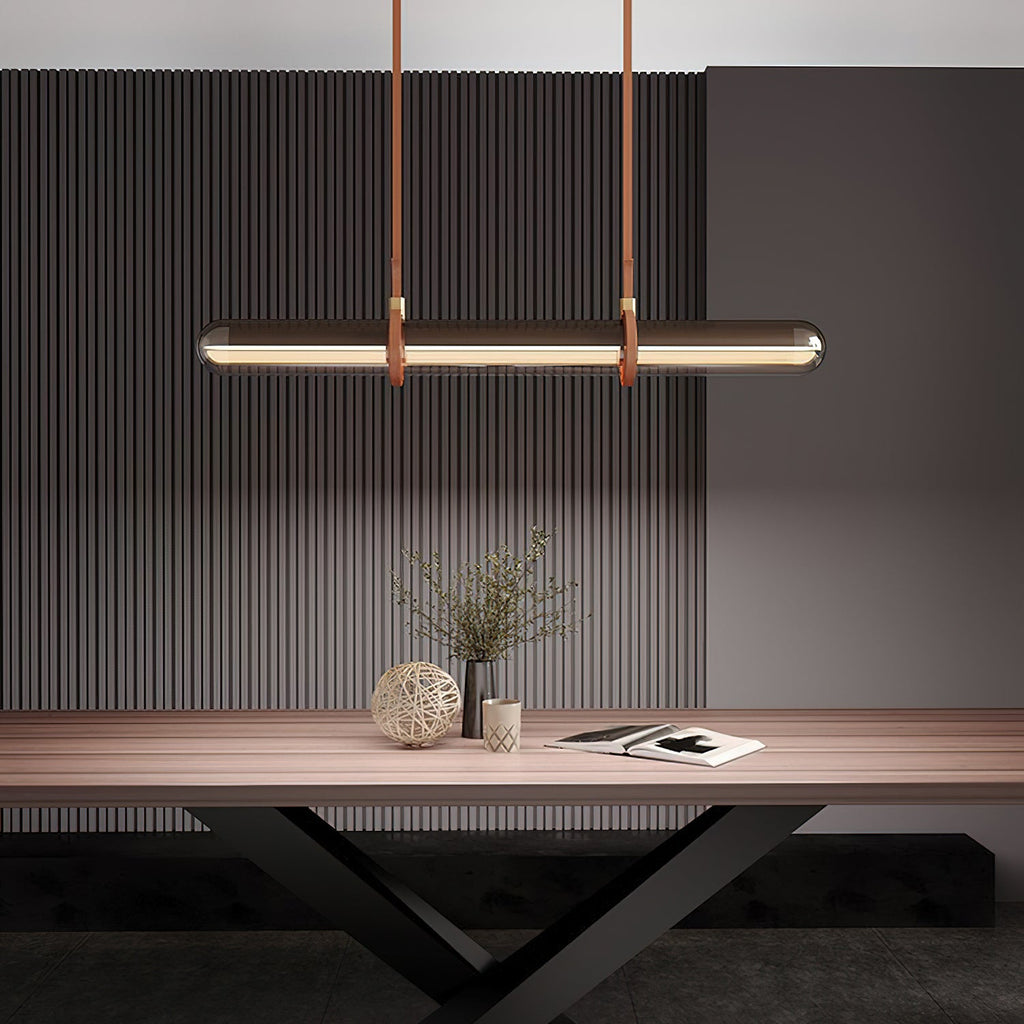 A Clérachic glass tube pendant glowing warm over a wooden table, with a plant in a vase and a book in front of a dark fluted wall.