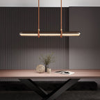 A Clérachic glass tube pendant glowing warm over a wooden table, with a plant in a vase and a book in front of a dark fluted wall.