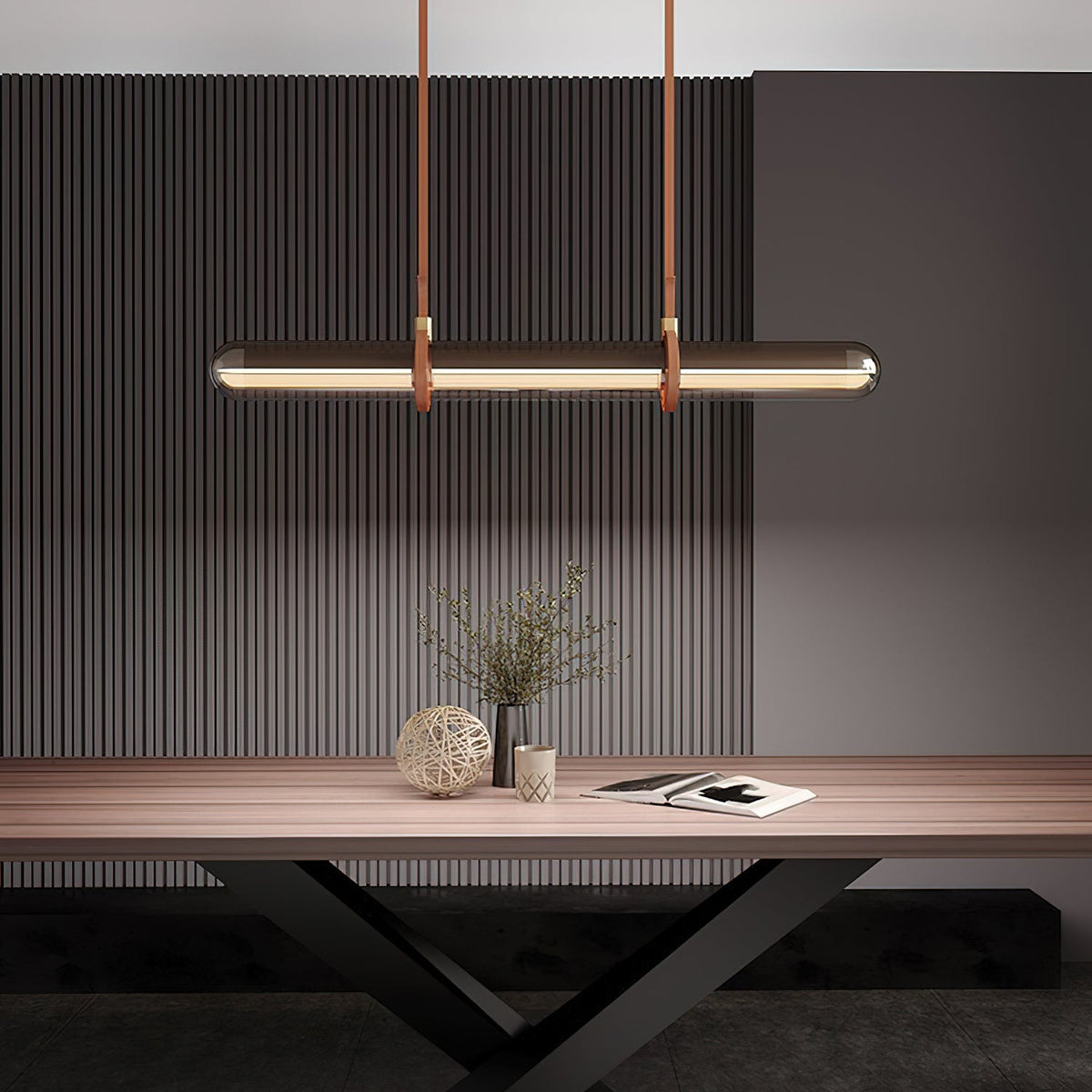A Clérachic glass tube pendant glowing warm over a wooden table, with a plant in a vase and a book in front of a dark fluted wall.