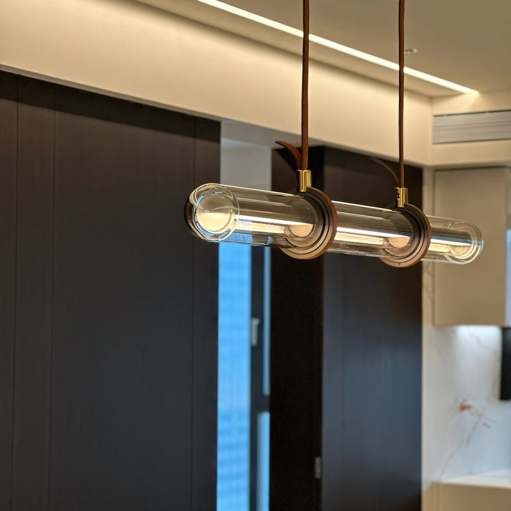Close view of the clear glass tube on this Clérachic pendant, with two brown leather loops and brass collars, in a dark-panelled room.