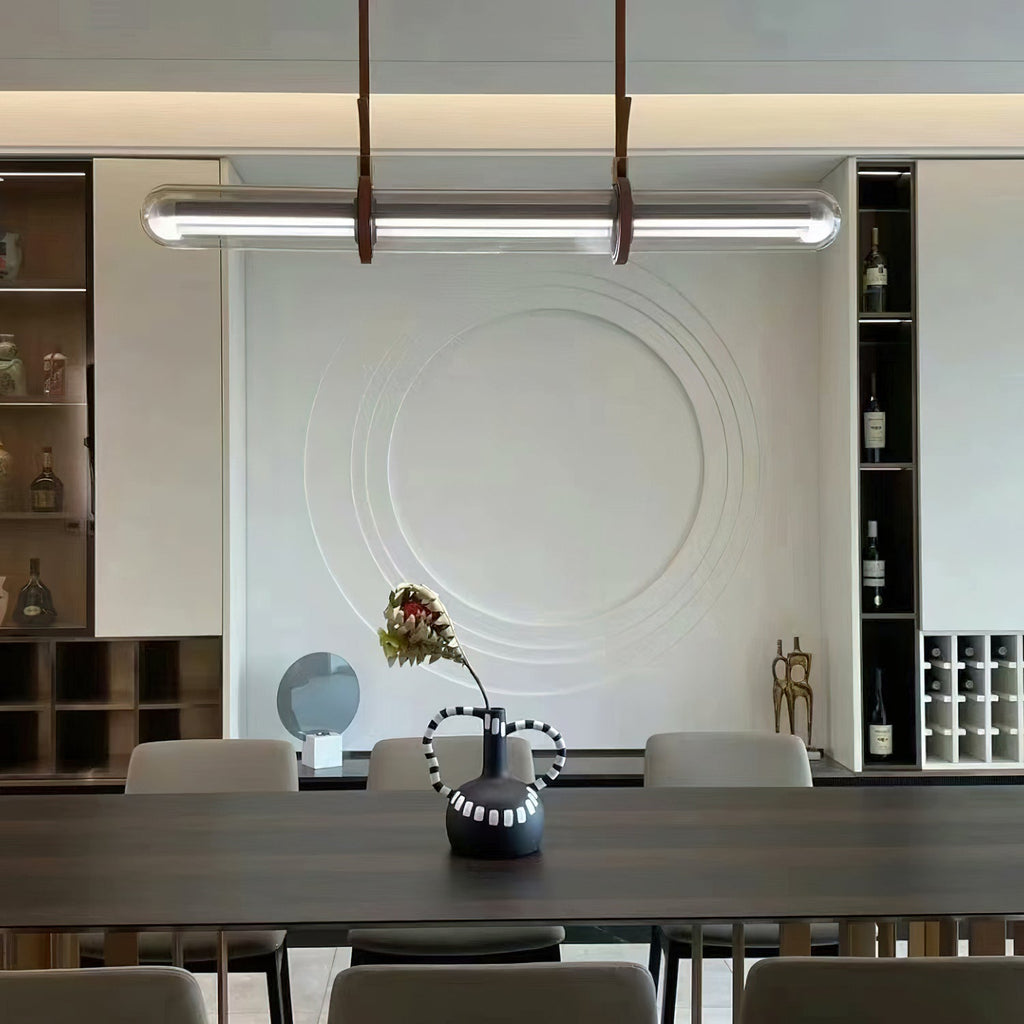 A Clérachic glass tube pendant over a dark dining table, in front of a white relief wall panel and shelves of bottles.